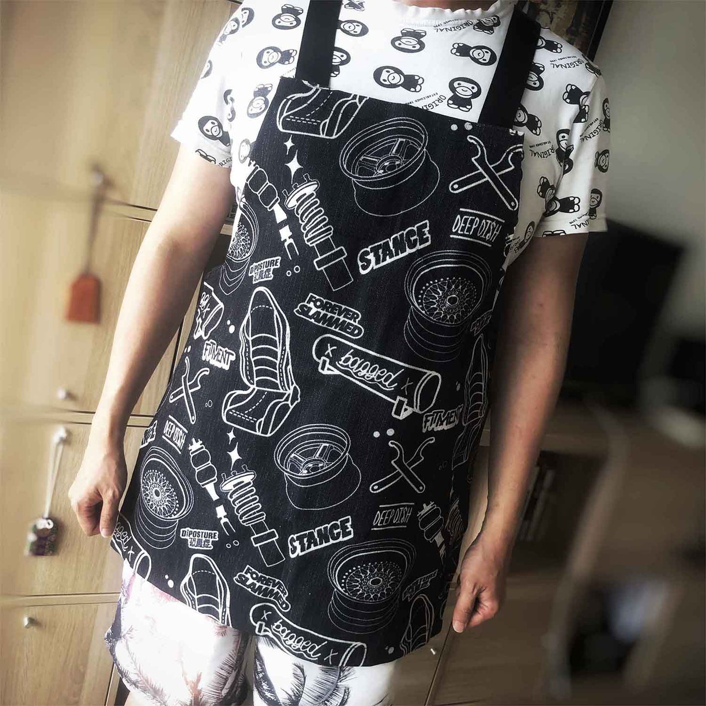 A man wearing a black JDM-themed apron with writings 'stance, forever slammed, fitment, bagged', standing in front of a cupboard