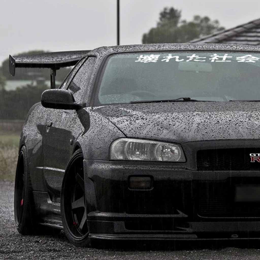 A white Broken Society car vinyl decal applied on a GTR's front window, and the car is parked in an open area while it is raining