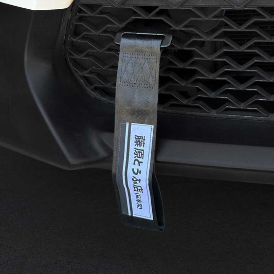 A black JDM tow strap with 'Fujiwara Tofu Shop' characters installed on the front mesh of a Toyota 86