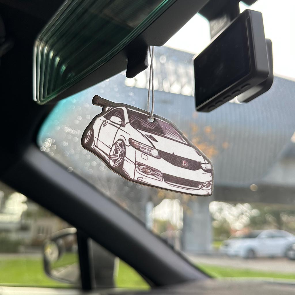 A Honda air freshener hung on a rearview mirror in a car with blurred building at the back