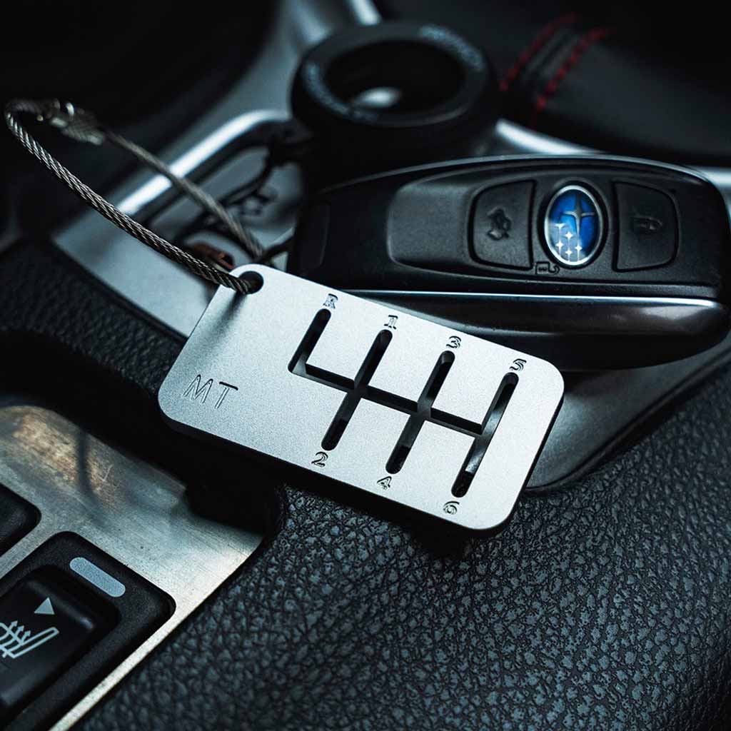 A six-speed manual shift keychain with matte silver finish placed in a car with a subaru car key