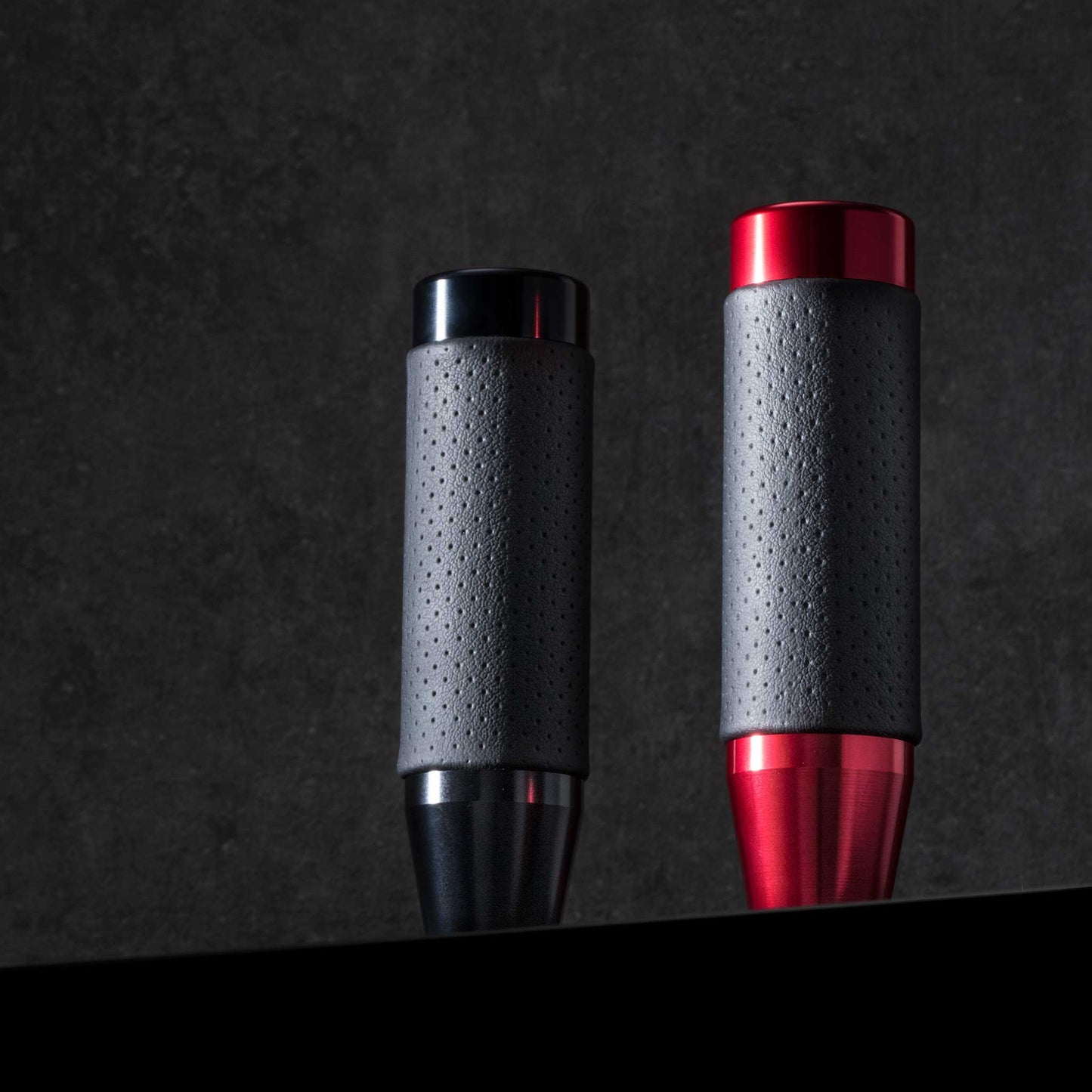 Two leather-grip shift knobs in different colours are standing on an black object and being overlooked, on a black background