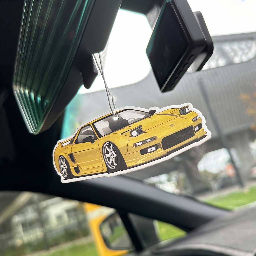A yellow Honda NSX air freshener hung on a rearview mirror in a car with blurred building at the back