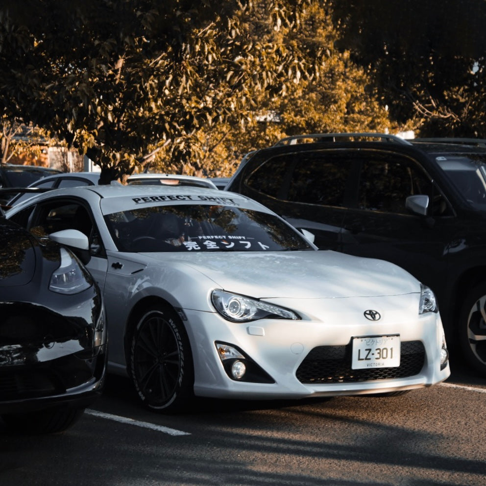 A white Toyota gt86 with Perfect Shift banner and decal parked in a public parking area in between two black cars