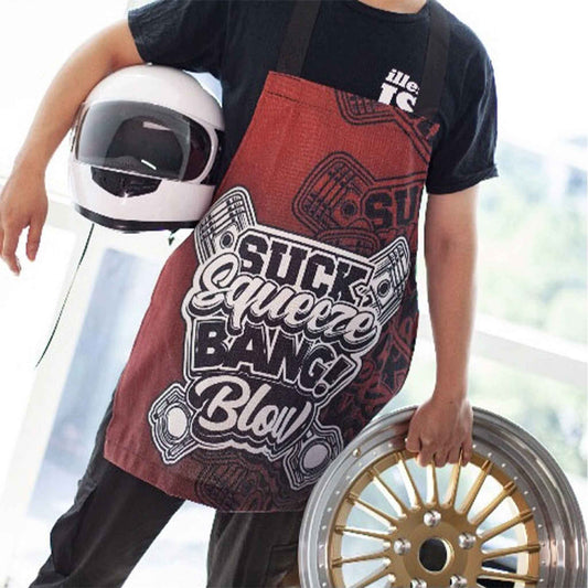 A man wearing a red JDM-themed apron with writings 'suck, squeeze, bang, blow' and holding a car wheel as well as a helmet, standing in front of a window