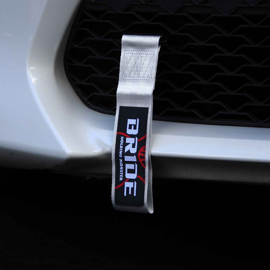 A silver BRIDE tow strap installed on a white car's front grille