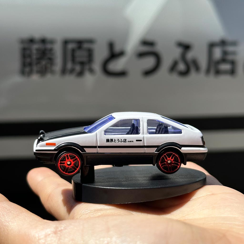 A hand holding an AE86 alloy car model in front of a real-life tofu 86