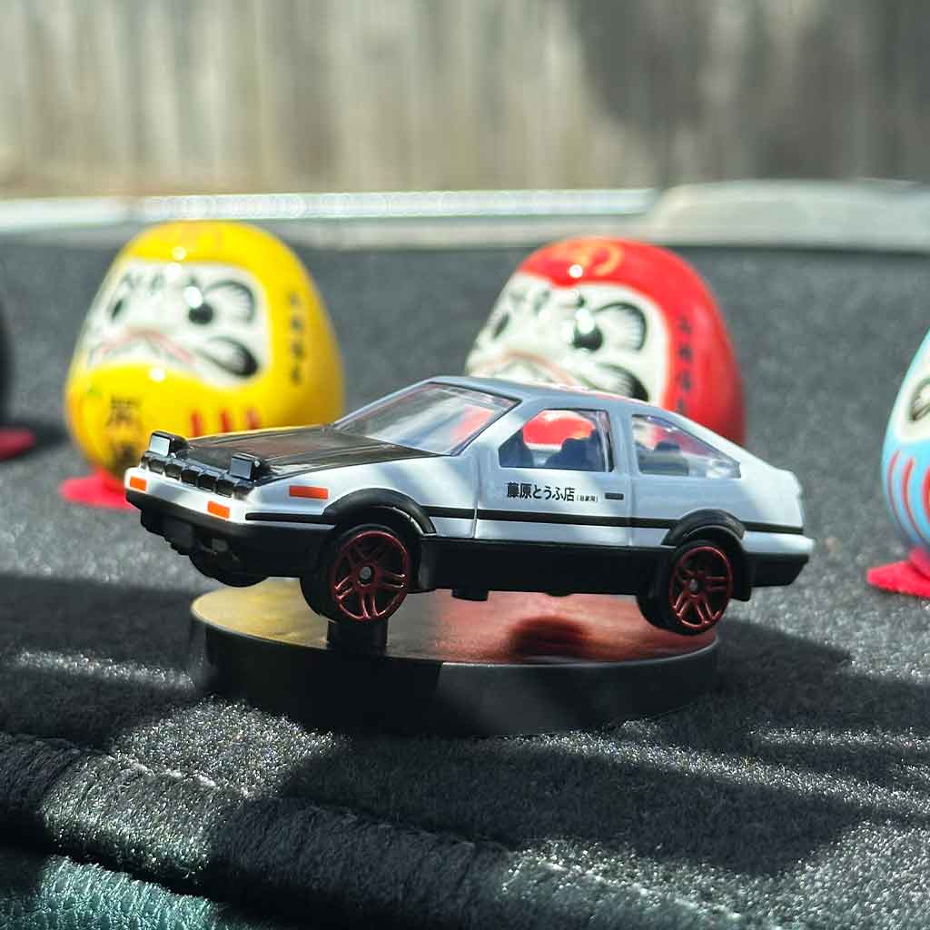 An AE86 car model drifter installed on a dashboard with some daruma porcelain statues behind 