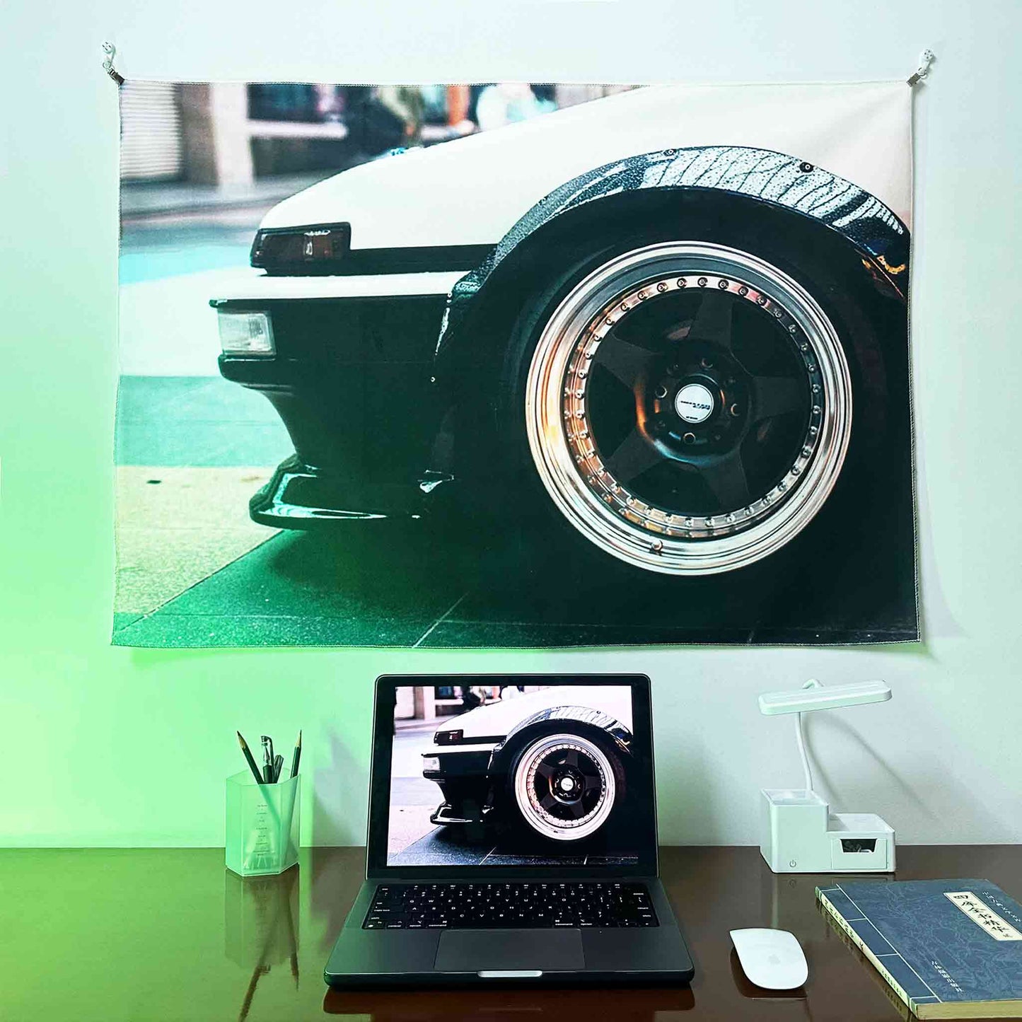 An AE86 artwork hung on a wall, with a laptop showing the same photo as the wall poster underneath, lit by green spotlight