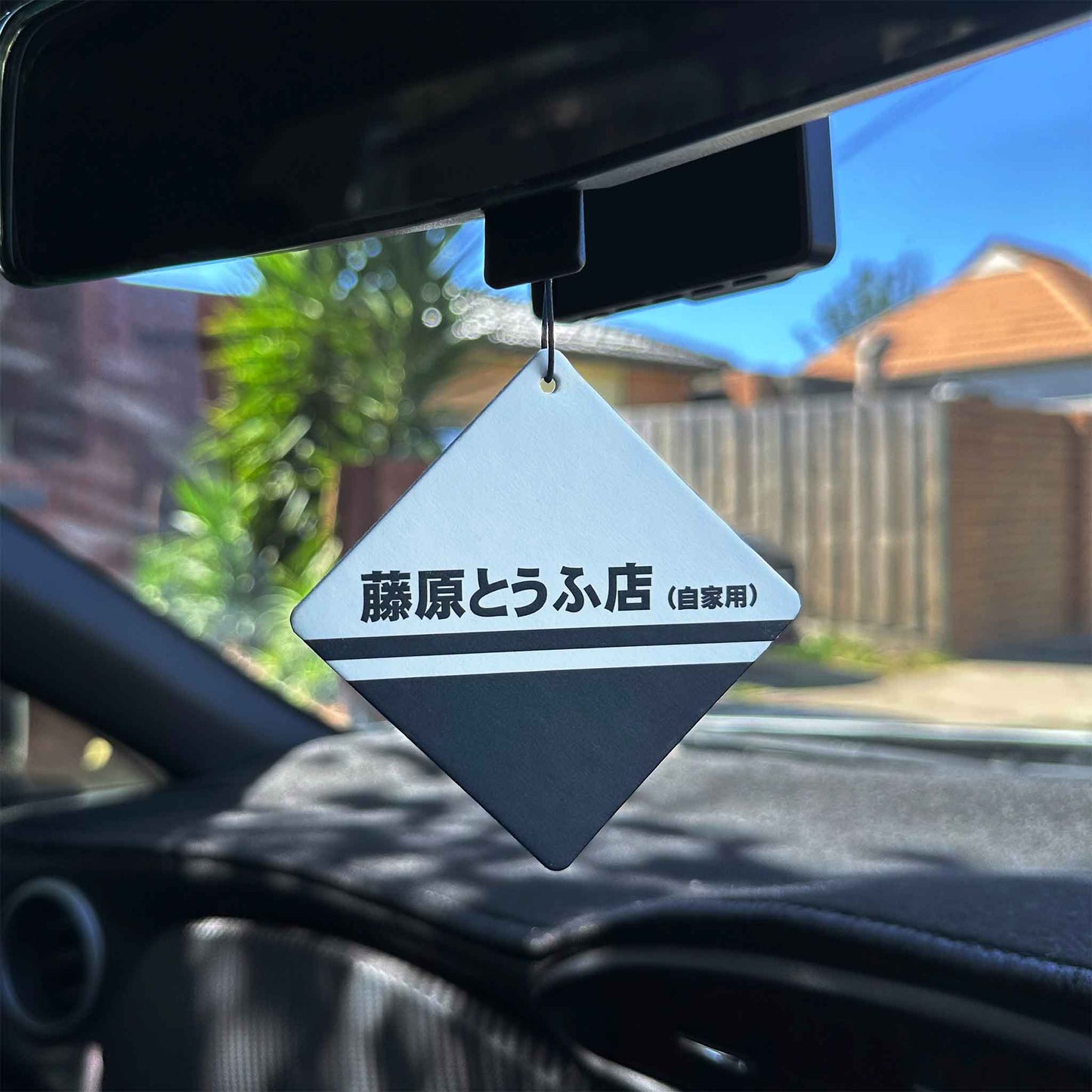 A Fujiwara-tofu-shop-themed rearview mirror air freshener hung in a car, with green tree and blue sky as background