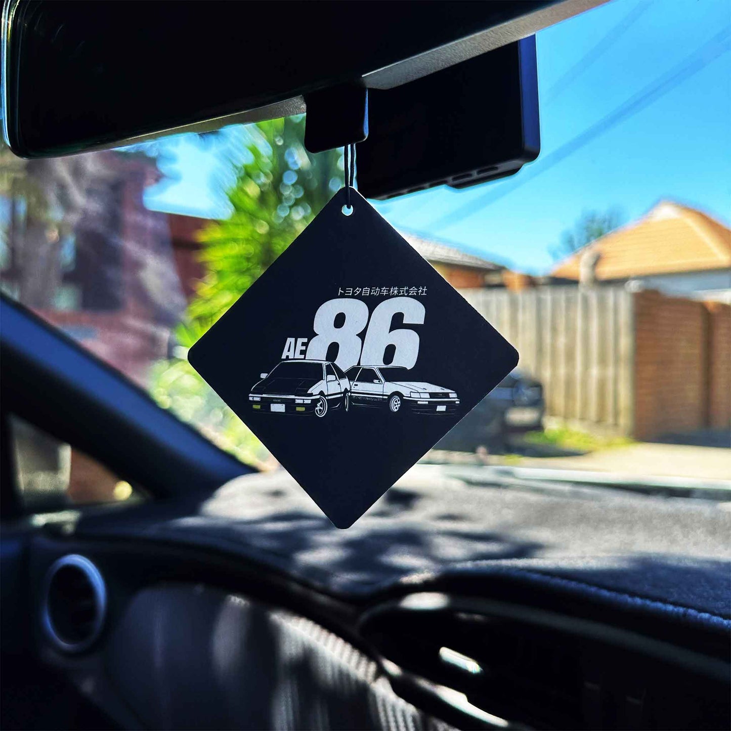 An AE86-themed rearview mirror air freshener hung in a car, with green tree and blue sky as background