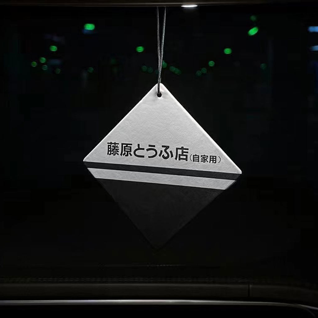 A Fujiwara-tofu-shop-themed rearview mirror air freshener hung in a car, with black background