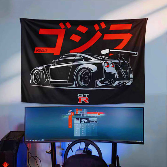 A GTR35 artwork hung on a wall, with a laptop screen showing a racing game and also a steering wheel simulator underneath, lit by natural light