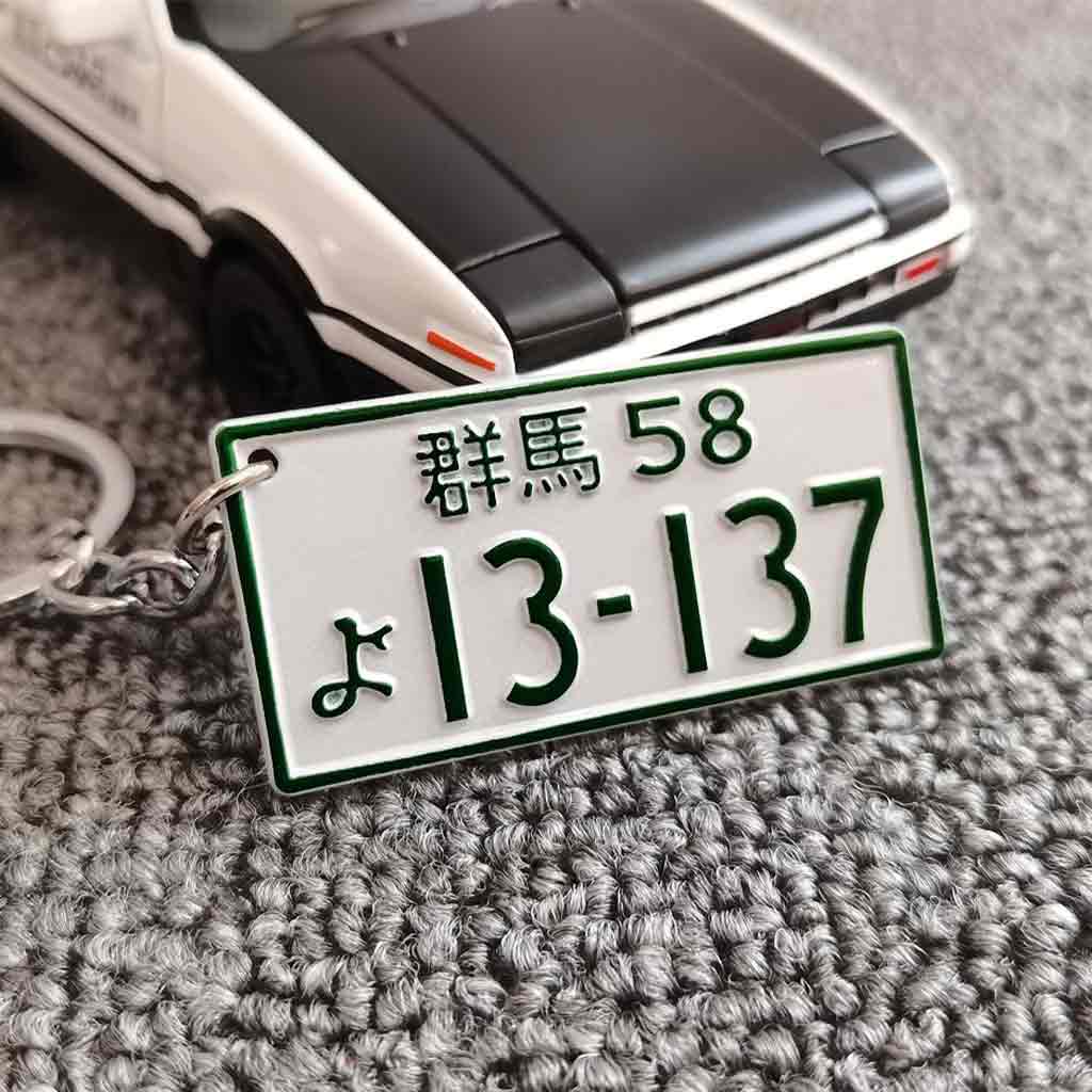 A metal keychain in front of an AE86 car model featuring the car number plate 'Gunma 13137'