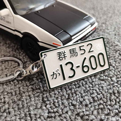 A metal keychain in front of an AE86 car model featuring the car number plate 'Gunma 13600'