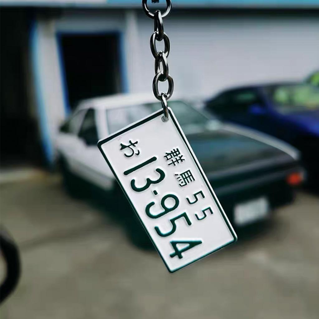 A metal keychain in front of a blurred AE86 featuring the car number plate 'Gunma 13954'