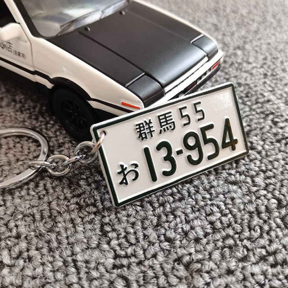 A metal keychain in front of an AE86 car model featuring the car number plate 'Gunma 13954'