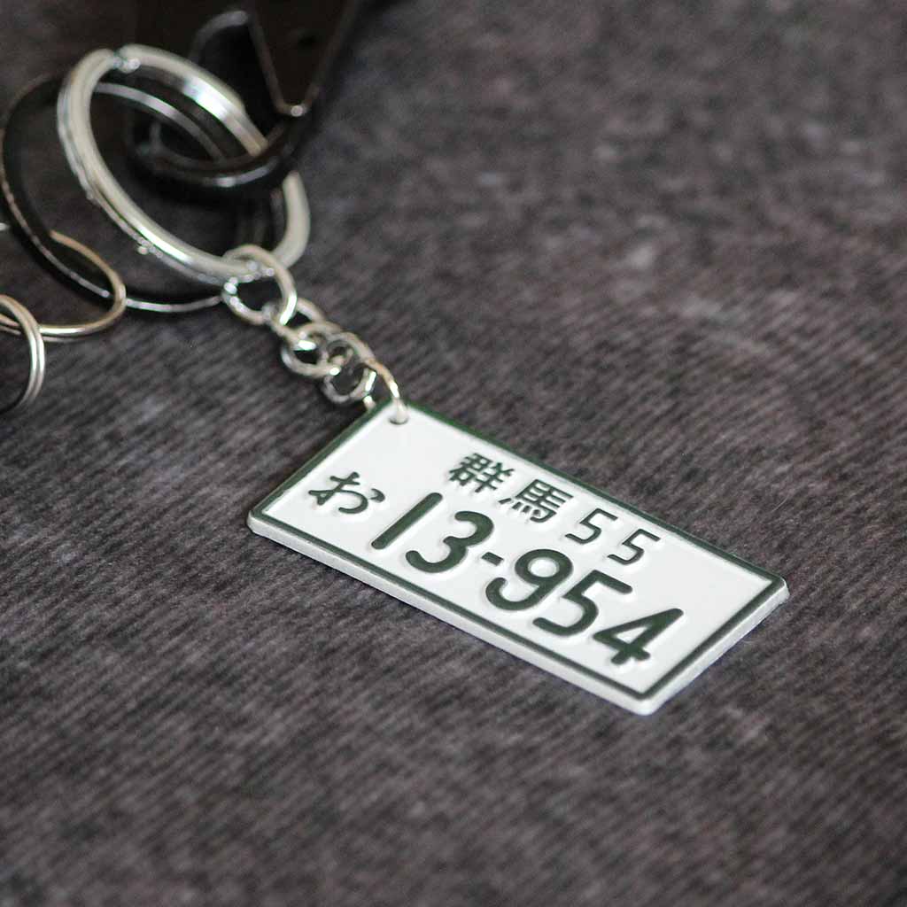 A white metal keychain with green characters 'Gunma55 13954' and outlines placed on a grey background
