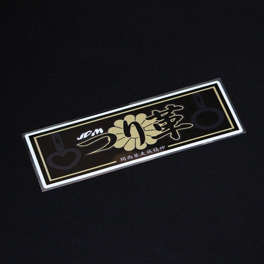 A gold JDM Tsurikawa slap sticker flat laid on a black background