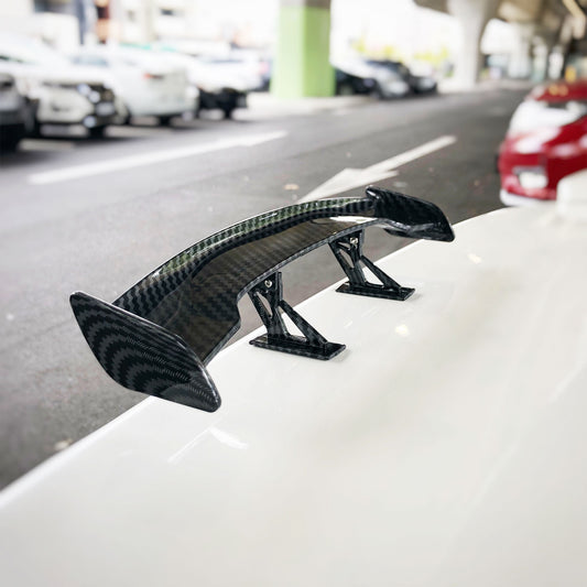 A mini spoiler made of carbon fibre installed on the back of a white car in a public parking area