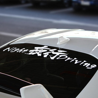 A white Night Driving car vinyl decal applied on a white car's rear window, and the car is parked in a public parking area