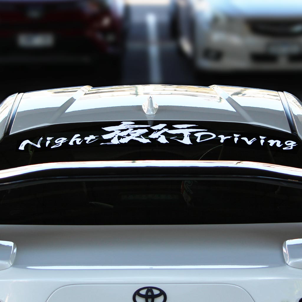 A white Night Driving car vinyl decal applied on a Toyota's rear window, and the car is parked in a public parking area