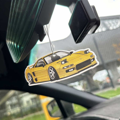 A yellow Honda NSX air freshener hung on a rearview mirror in a car with blurred building at the back