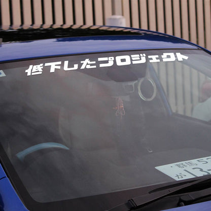 A white Lowered Project car vinyl decal applied on a blue car's front window with wooden fences behind the car