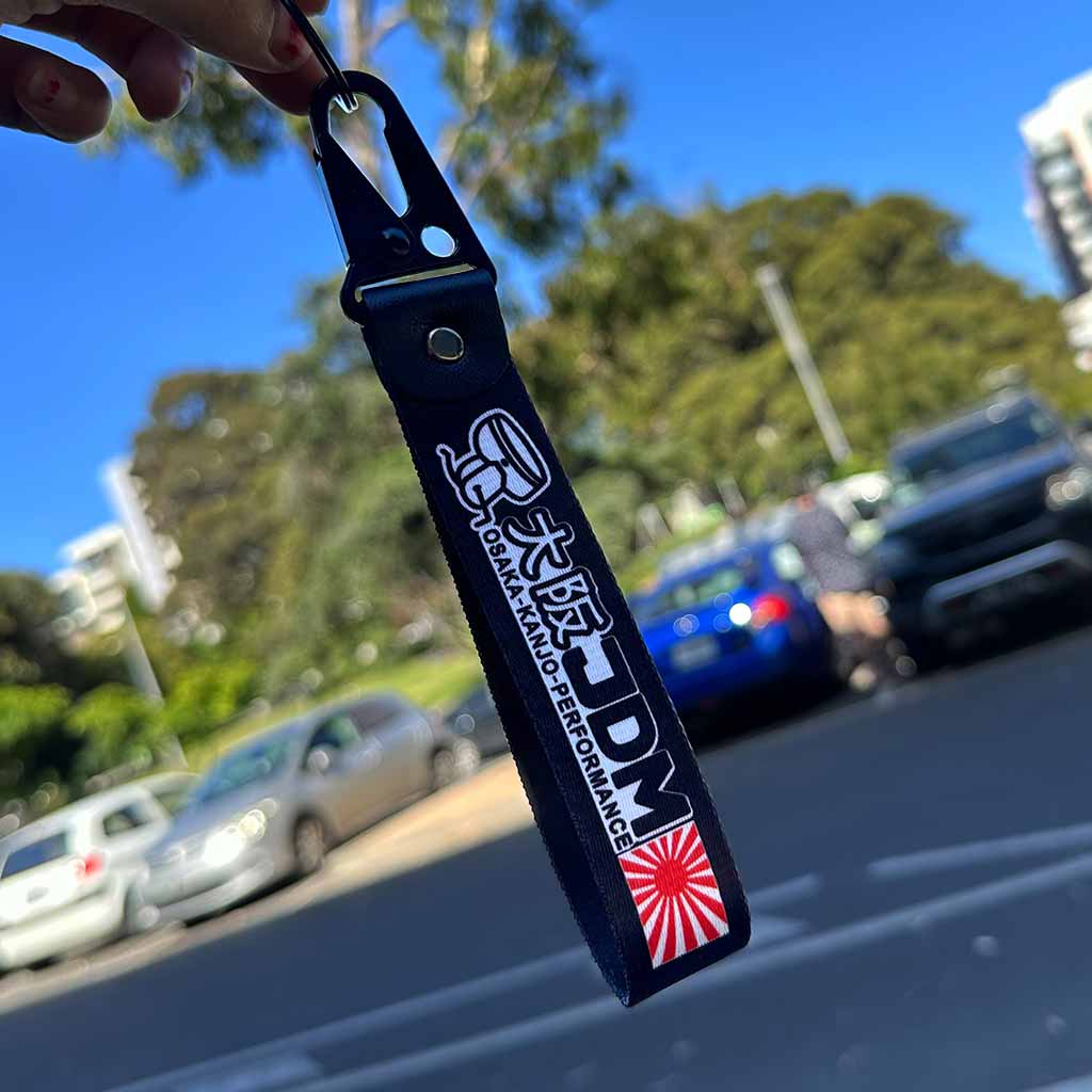 Hand holding a Osaka Kanjo Performance lanyard in front of cars and blue sky