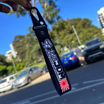 Hand holding a Osaka Kanjo Performance lanyard in front of cars and blue sky