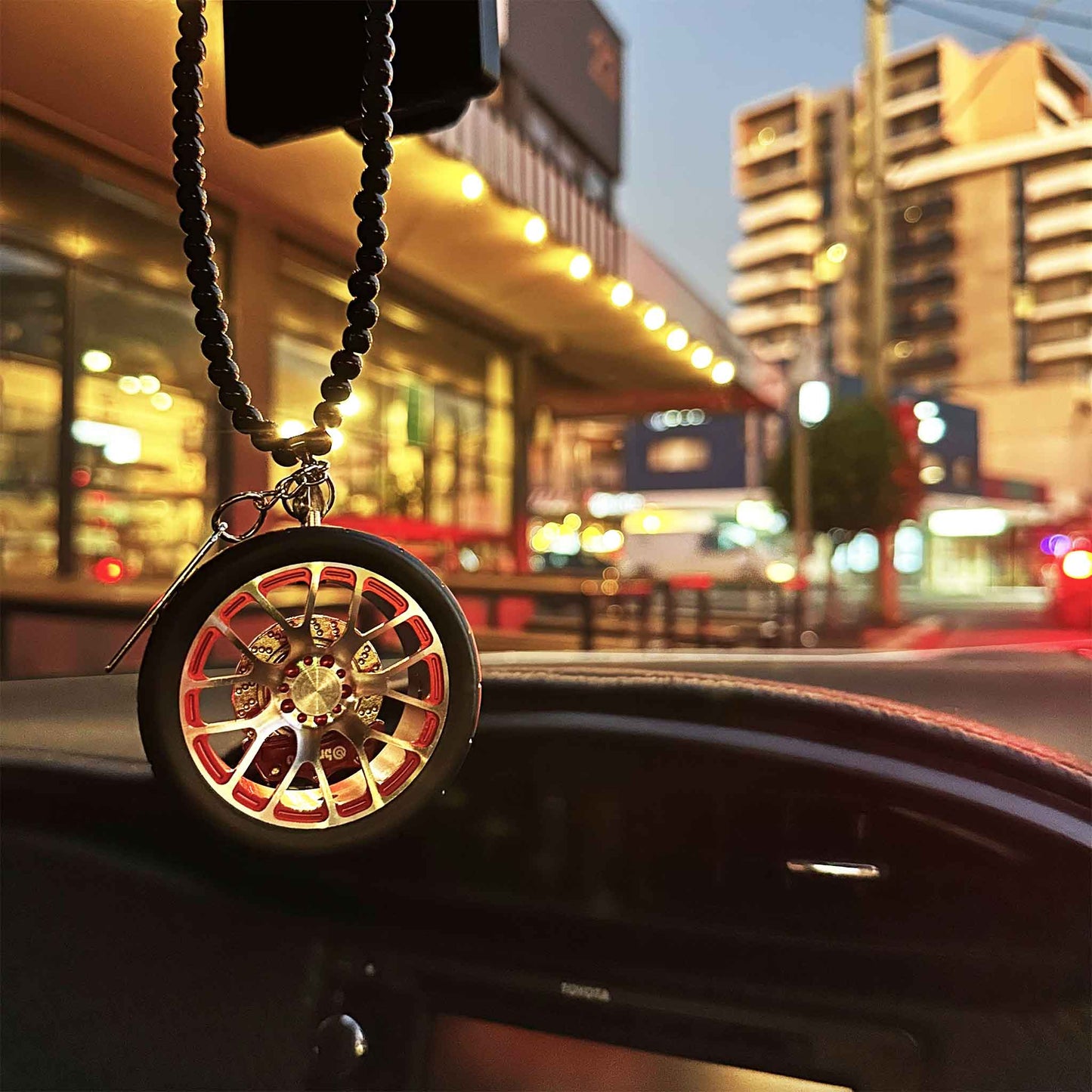 A red BBS air-freshener hung on a car's rear mirror, with well-lit but blurred street views at the background