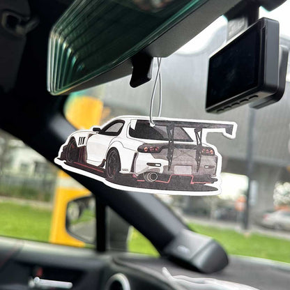 An RX7 air-freshener hung on a rearview mirror with dash cam and blurred street views at the back