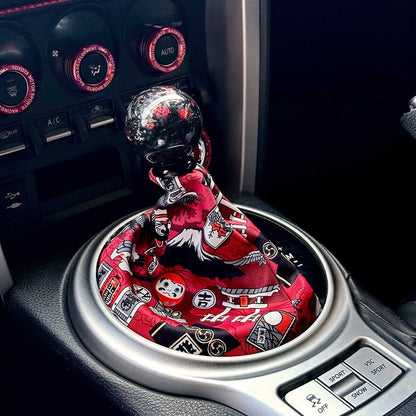 A cute JDM shift boot featuring daruma and other Japanese elements installed in an automatic Toyota 86, with a red carbon fibre shift knob on top