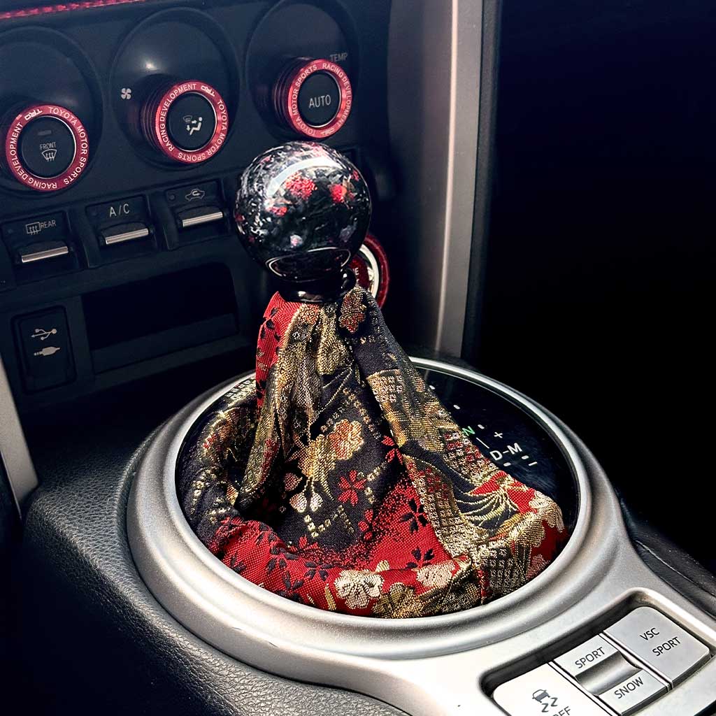 A cool JDM shift boot featuring golden and red cherry blossoms and Japanese fans installed in an automatic Toyota 86, with a red carbon fibre shift knob on top