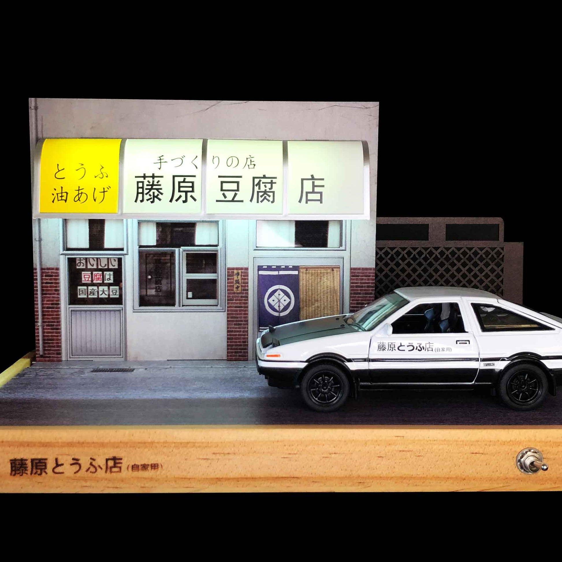 Front of a Fujiwara tofu shop and side of an AE86 car model both on a wooden base