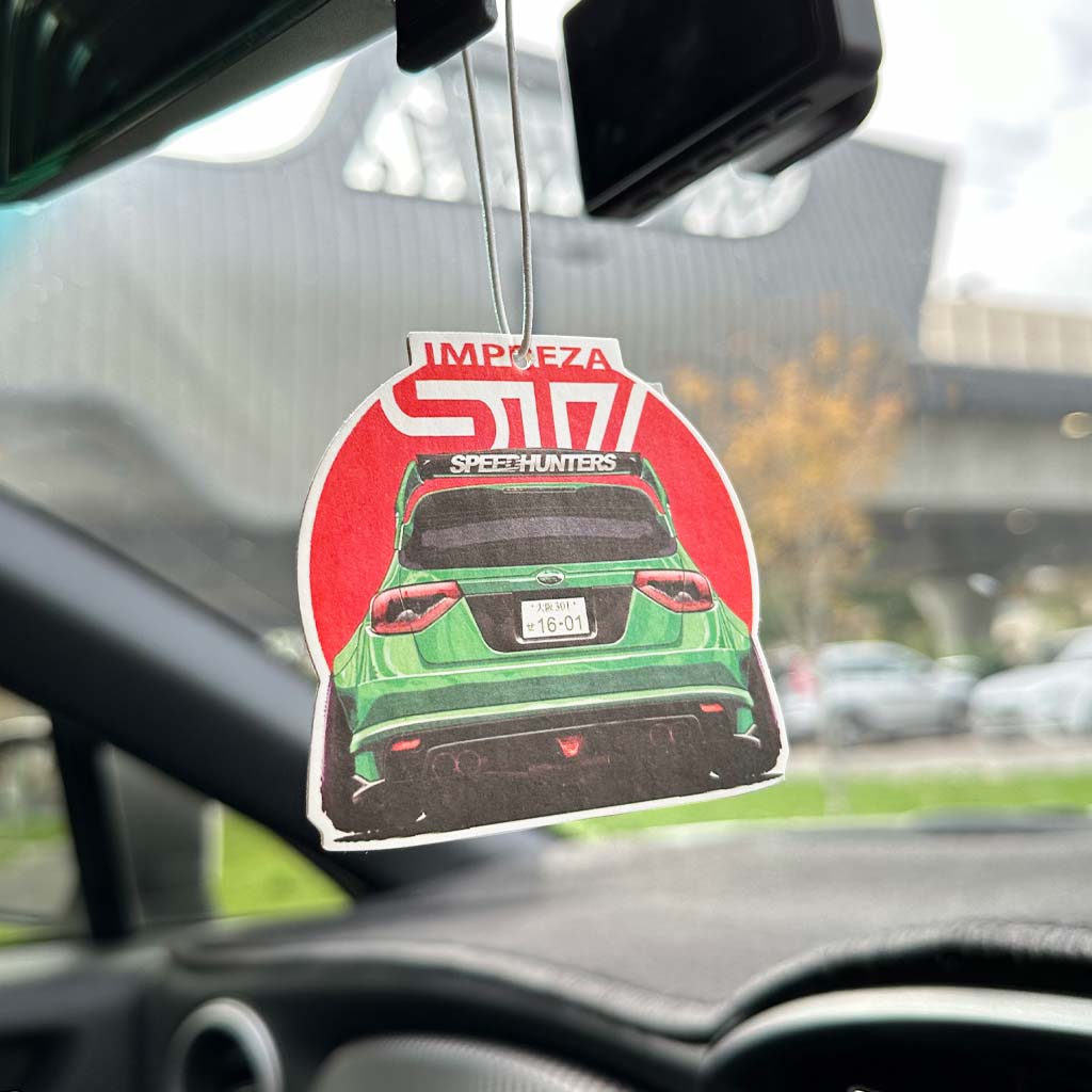 A Impreza STI air-freshener hung on a rearview mirror in a car with blurred building at the back