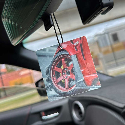 An air freshener highlighting a red car with red Volk Racing wheel is hung on a car's rearview mirror