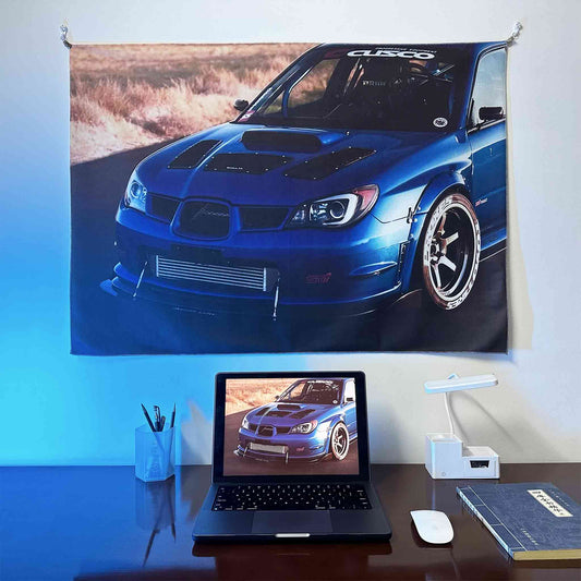 A rally blue Subaru Sti artwork hung on a wall, with a laptop showing the same photo as the wall poster underneath, lit by blue spotlight
